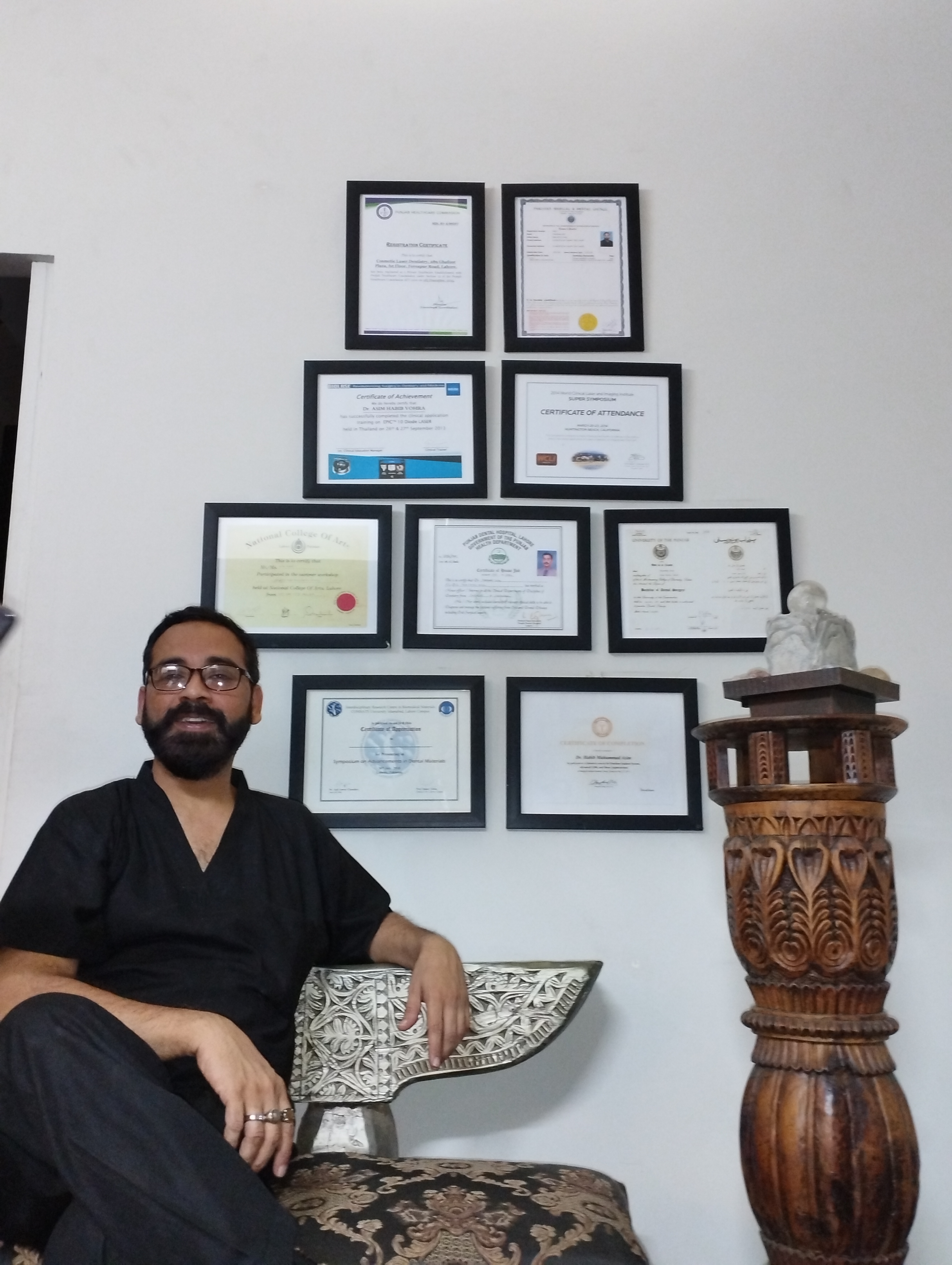 Dr. M. Asim Habib Vohra seated beneath his framed dental certificates